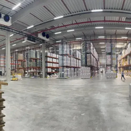 Großer Lagerraum mit Regalen voll mit Kisten und Waren, leerer Boden, Person in der Ferne.